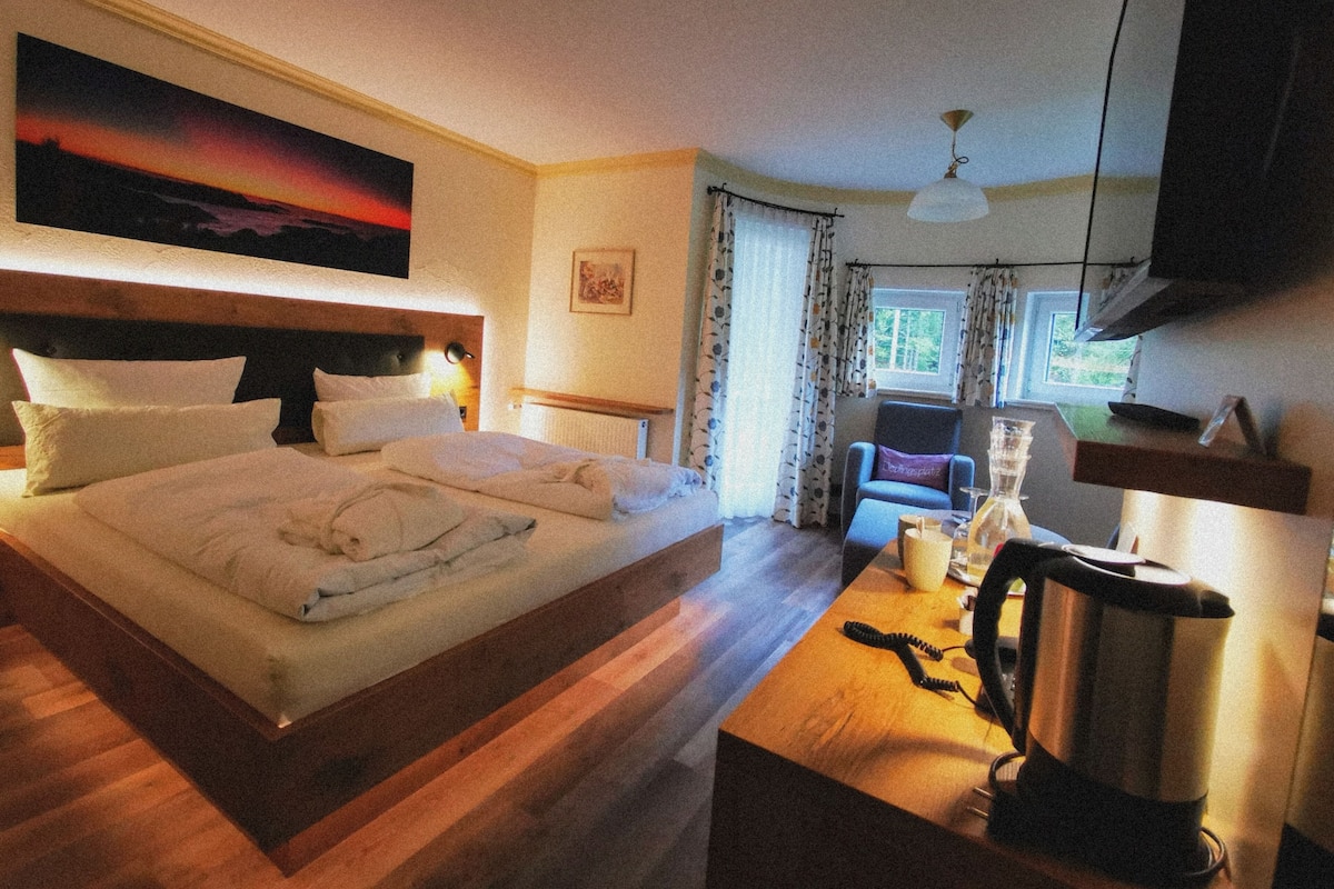 A hotel room with a large bed, a desk with a kettle, a chair, and a television.