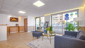 A modern hotel lobby with a grey sofa, a round coffee table, a blue ottoman, a large window with a view of the street, and a painting on the wall. - Hotel Parisien (Menton)