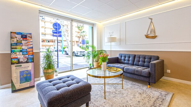 A modern lobby with a sofa, ottoman, and coffee table, a large window with a view of the street, and a bulletin board with various notices. - Hotel Parisien (Menton)