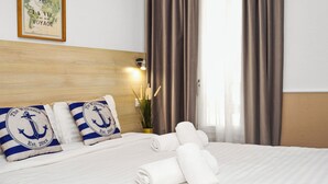A neatly made bed with rolled white towels, two pillows with nautical designs, a framed map on the wall, and a window with curtains. - Hotel Parisien (Menton)