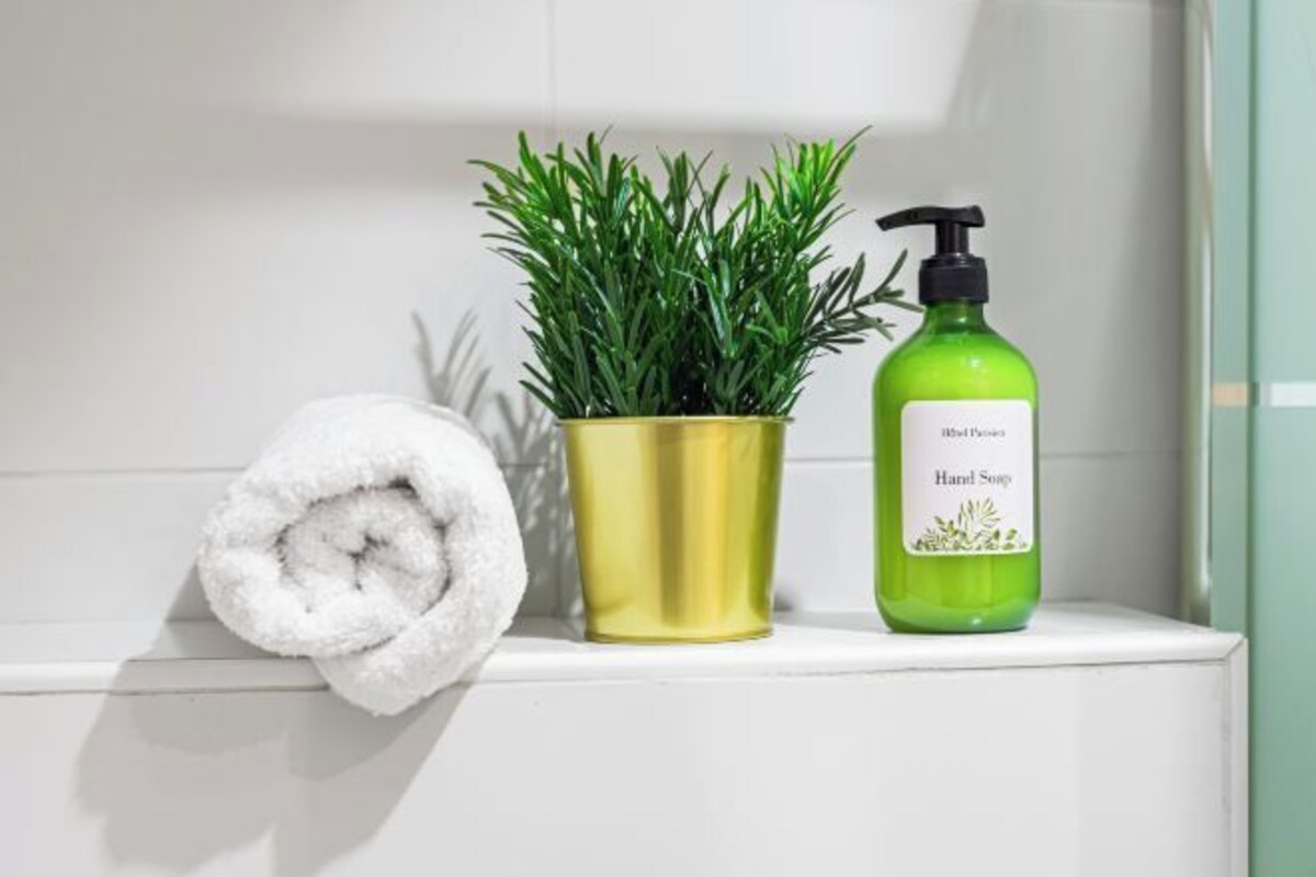 A bathroom with a white towel, a gold plant pot, and a green bottle of hand soap.