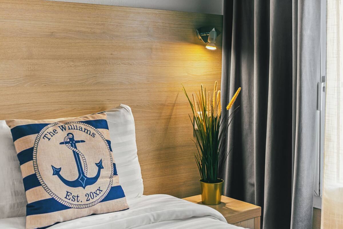 A neatly made bed with a pillow featuring an anchor design, a wooden headboard, a bedside lamp, and a potted plant.