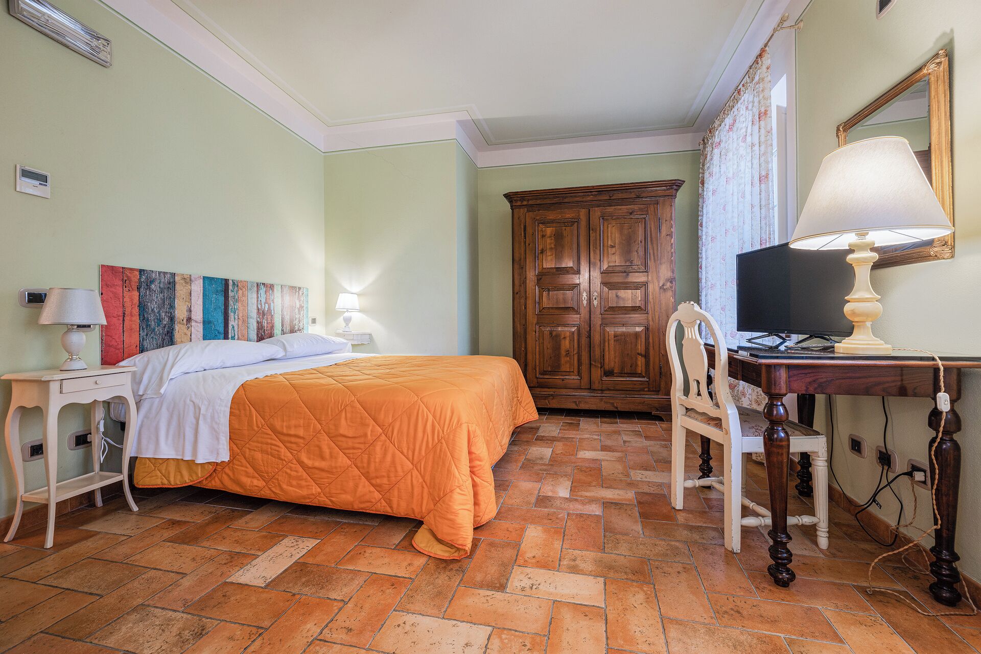 A bedroom with a bed, a desk with a lamp, a chair, a wardrobe, and a television.