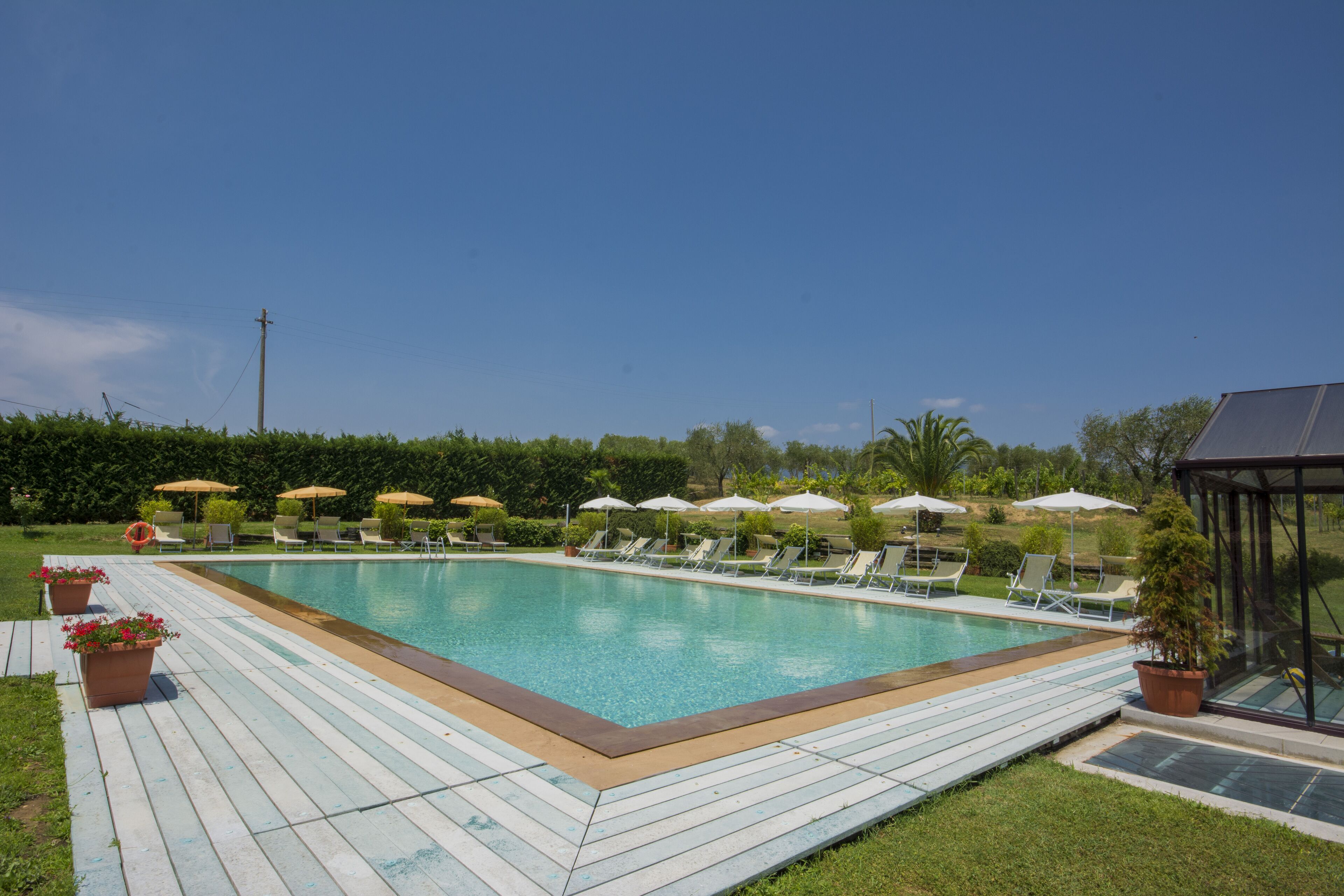 A rectangular swimming pool with a wooden deck, surrounded by lounge chairs and umbrellas, and a glass-enclosed structure to the right.