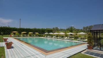 A rectangular swimming pool with a wooden deck, surrounded by lounge chairs and umbrellas, and a glass-enclosed structure to the right.