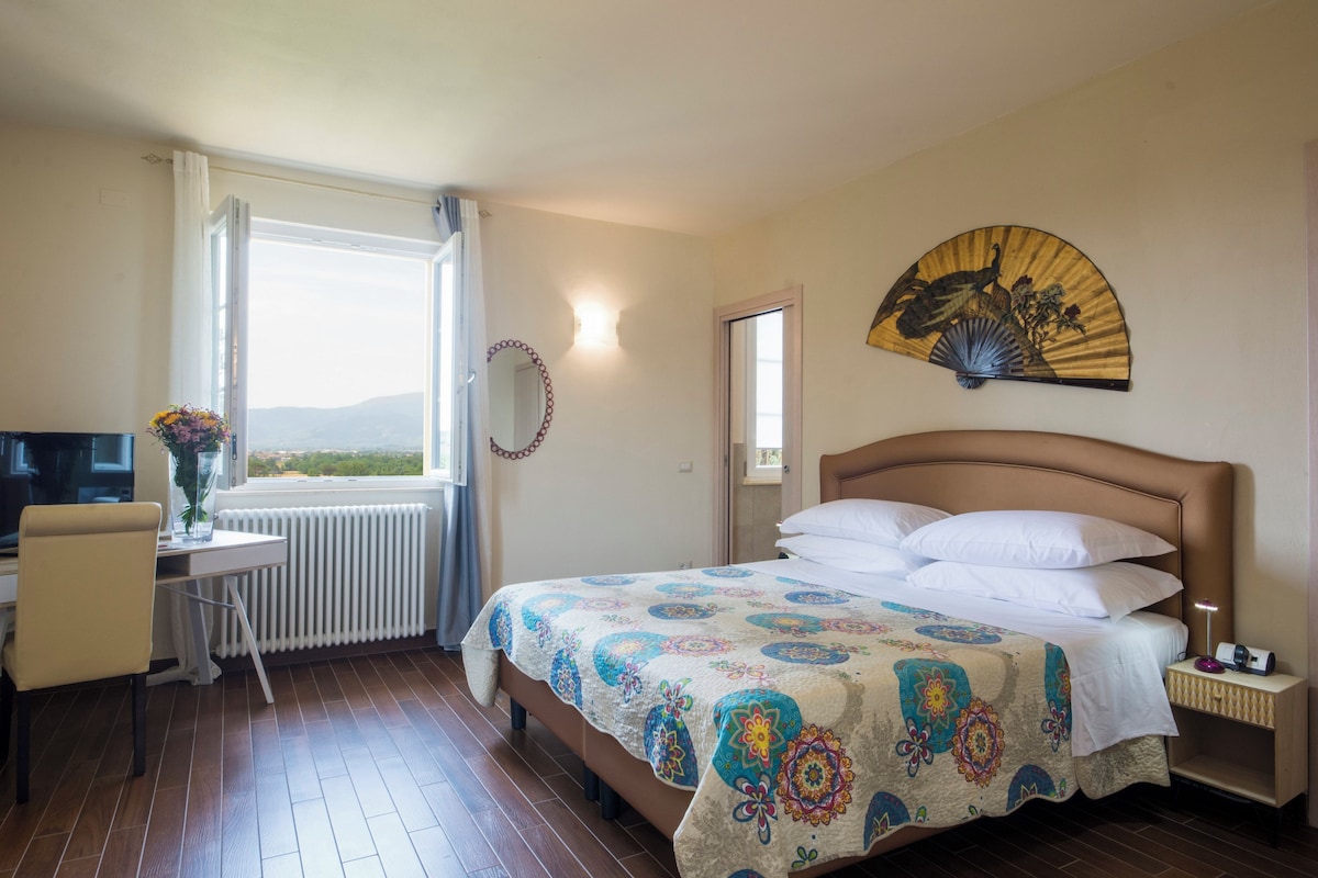 Une chambre d’hôtel comprenant un lit, un bureau avec une chaise, une télévision, une fenêtre avec vue et un ventilateur décoratif fixé au mur.