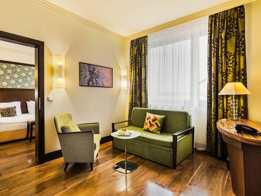 Ein Hotelzimmer mit Bett, Sofa, Sessel, kleinem Tisch und Schreibtisch mit Lampe.