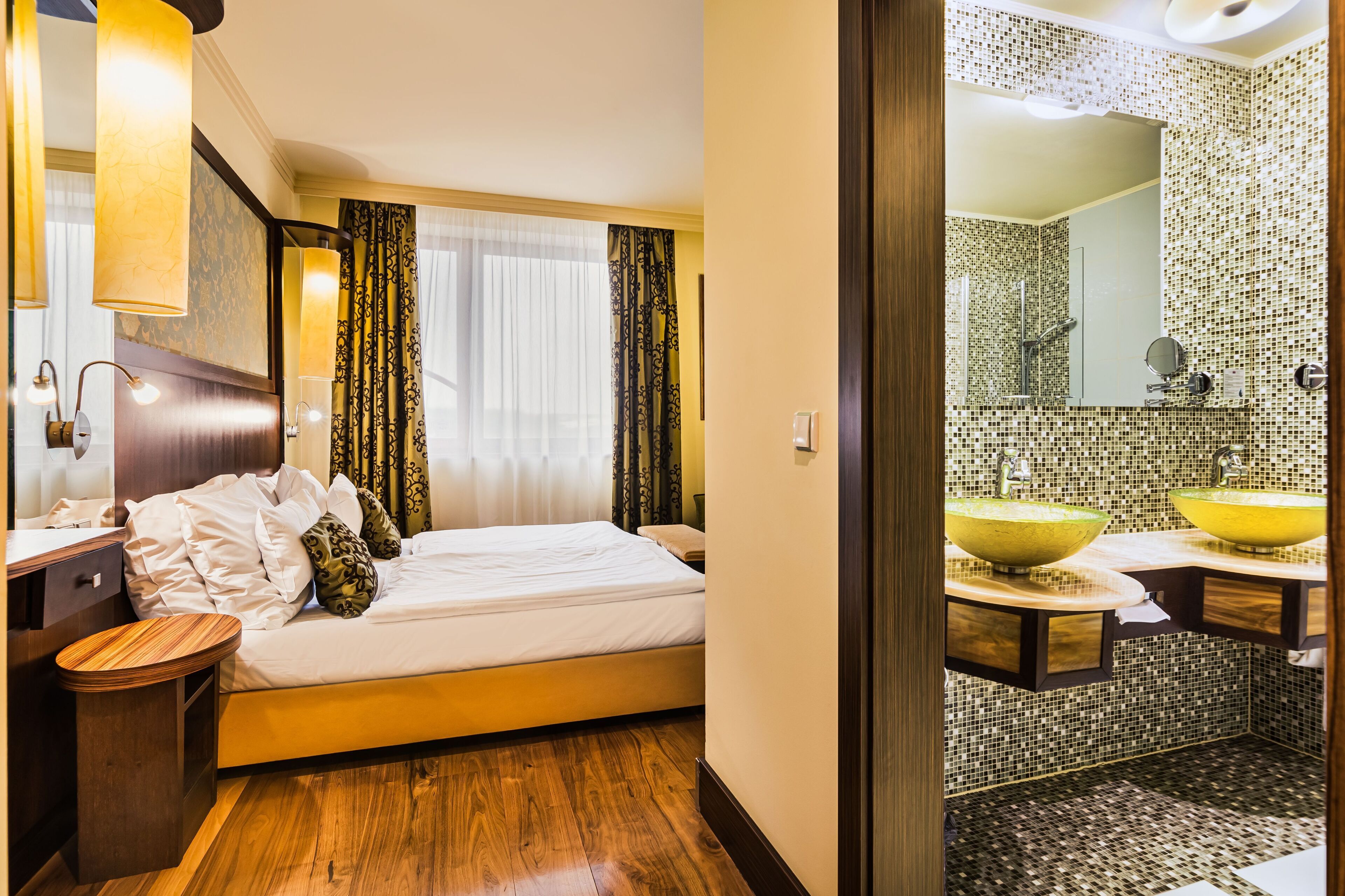 A hotel room with a bed, bedside table, lamp, curtains, and a bathroom with a sink and mirror.