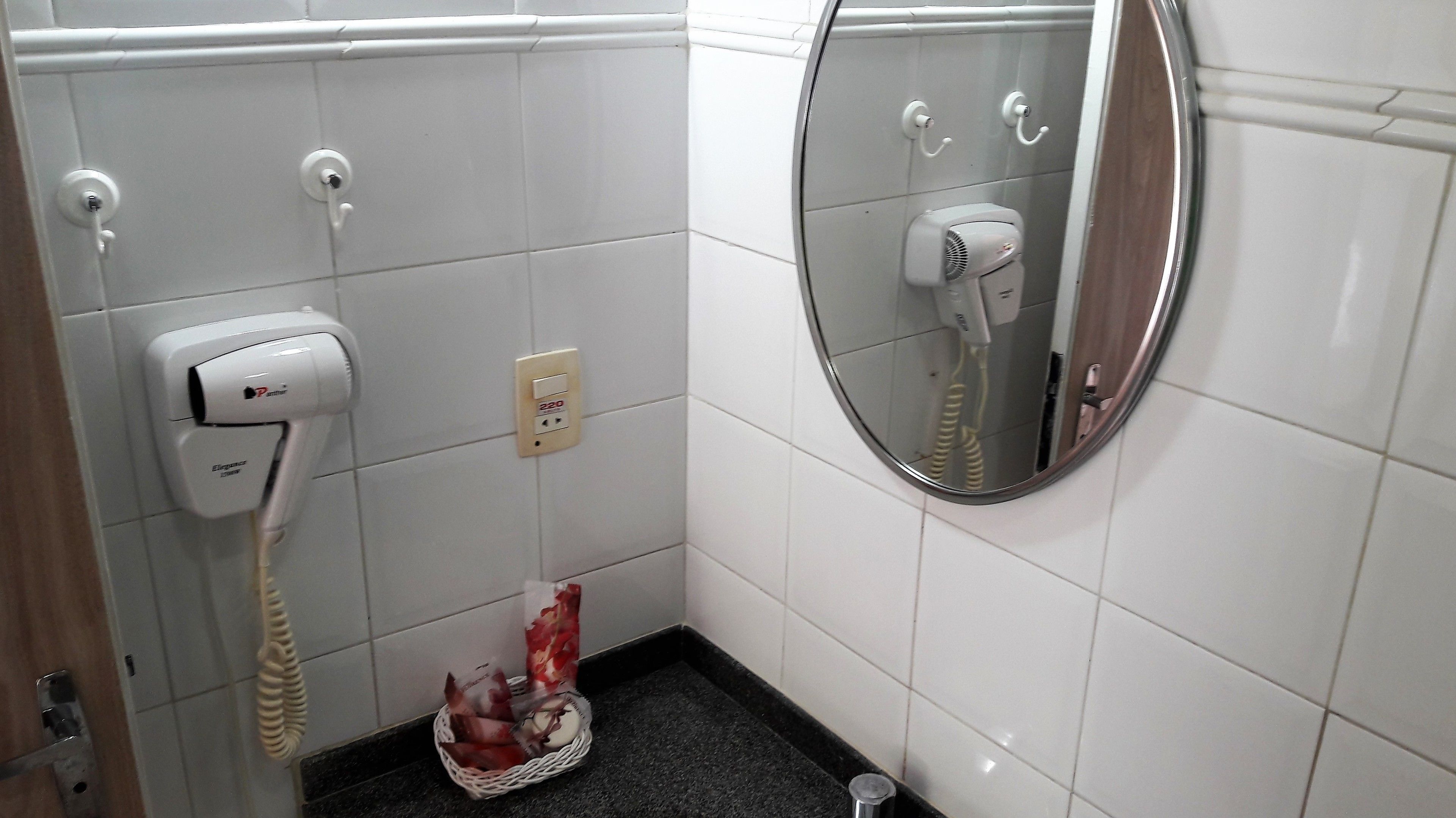 Ett badrum med vita kakelväggar, en handtork, en hårtork och en korg med toalettartiklar.