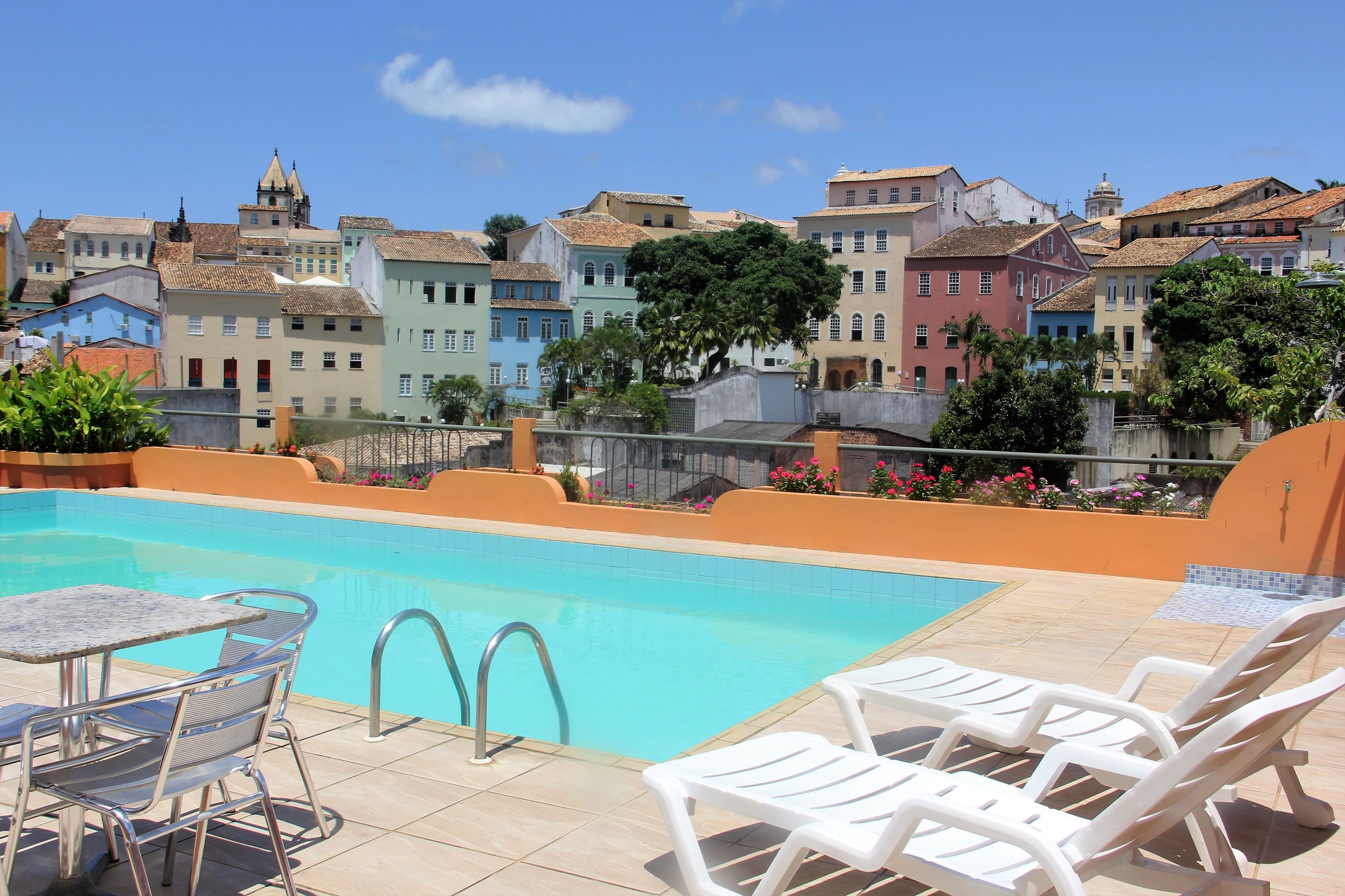 A rooftop pool area with a view of colorful buildings, a table with chairs, and a small garden with flowers.