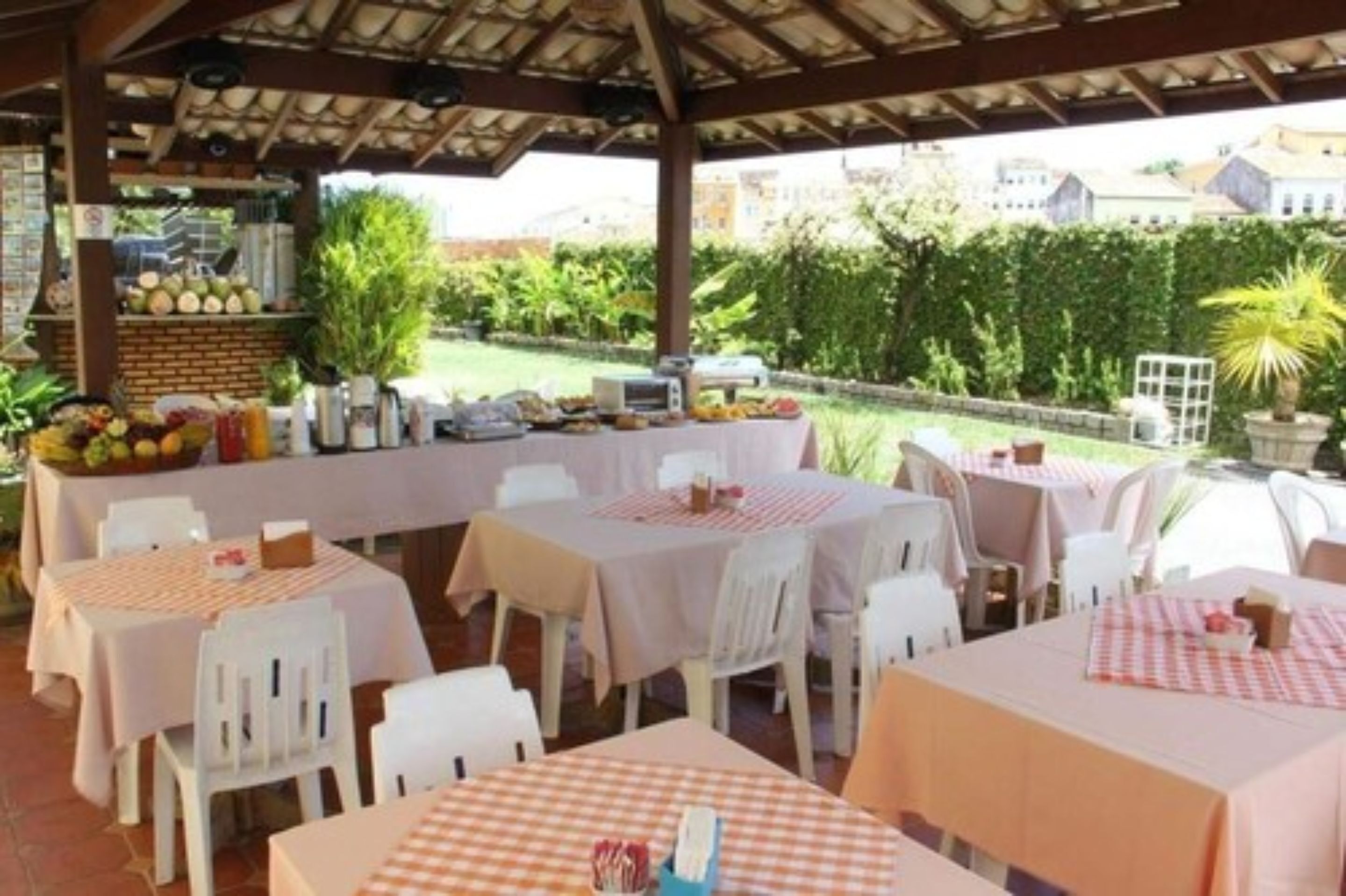 An outdoor dining area with tables covered in checkered tablecloths, white chairs, and a variety of fruits and vegetables displayed on a stand.