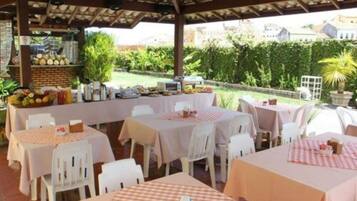 An outdoor dining area with tables covered in checkered tablecloths, white chairs, and a variety of fruits and vegetables displayed on a stand.