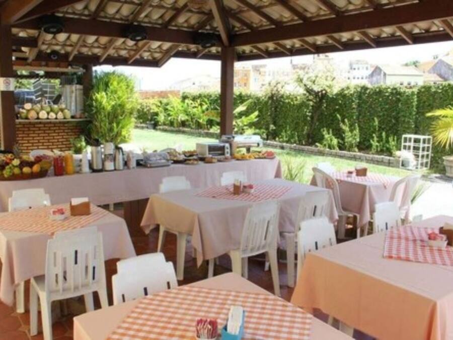 An outdoor dining area with tables covered in checkered tablecloths, white chairs, and a variety of fruits and vegetables displayed on a stand.