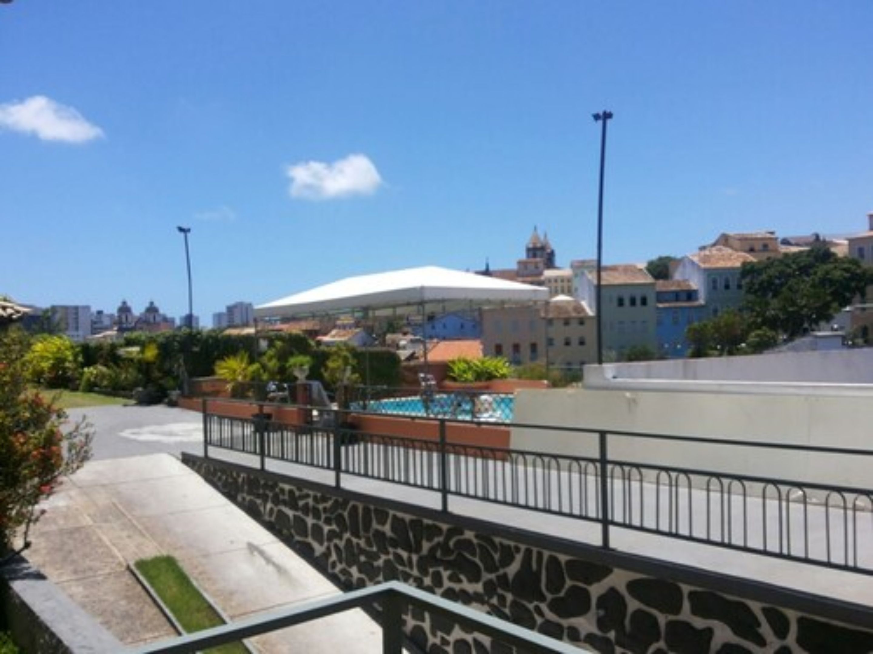 Vista de una zona de piscina con muro de piedra y barandilla metálica, rodeada de edificios y vegetación.