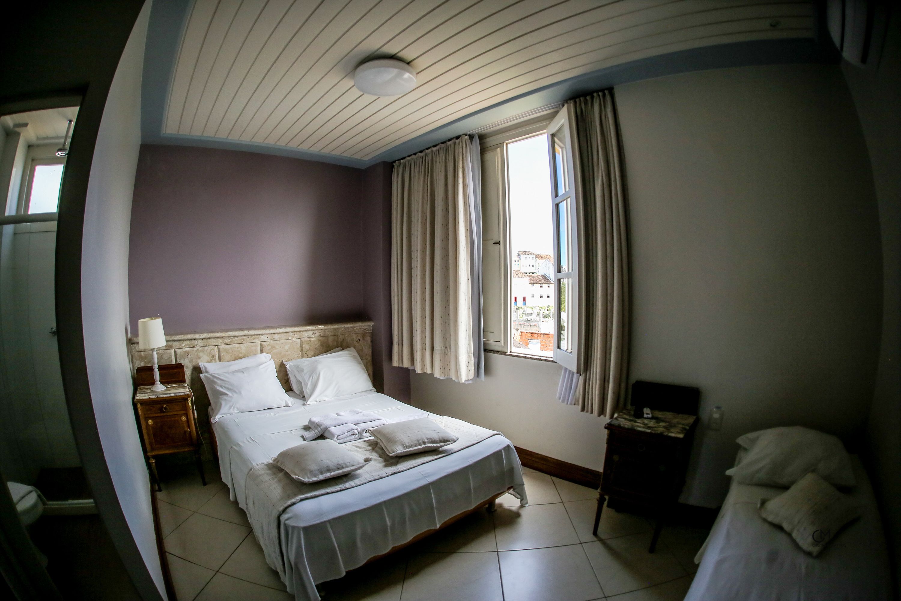 A hotel room with a bed, a nightstand, a window with curtains, and a view of buildings outside.