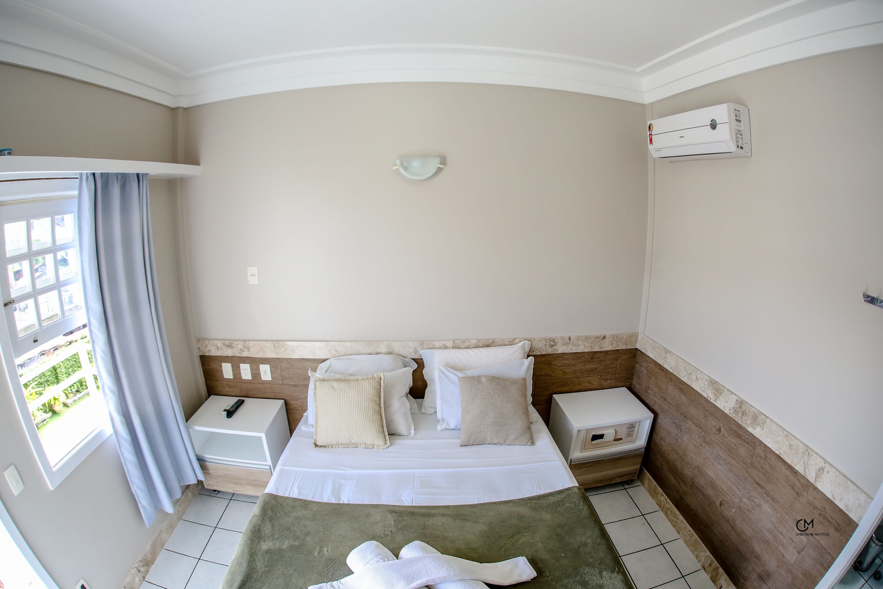 Habitación de hotel con cama, mesitas de noche, ventana con cortinas, equipo de aire acondicionado y vistas a zonas verdes.