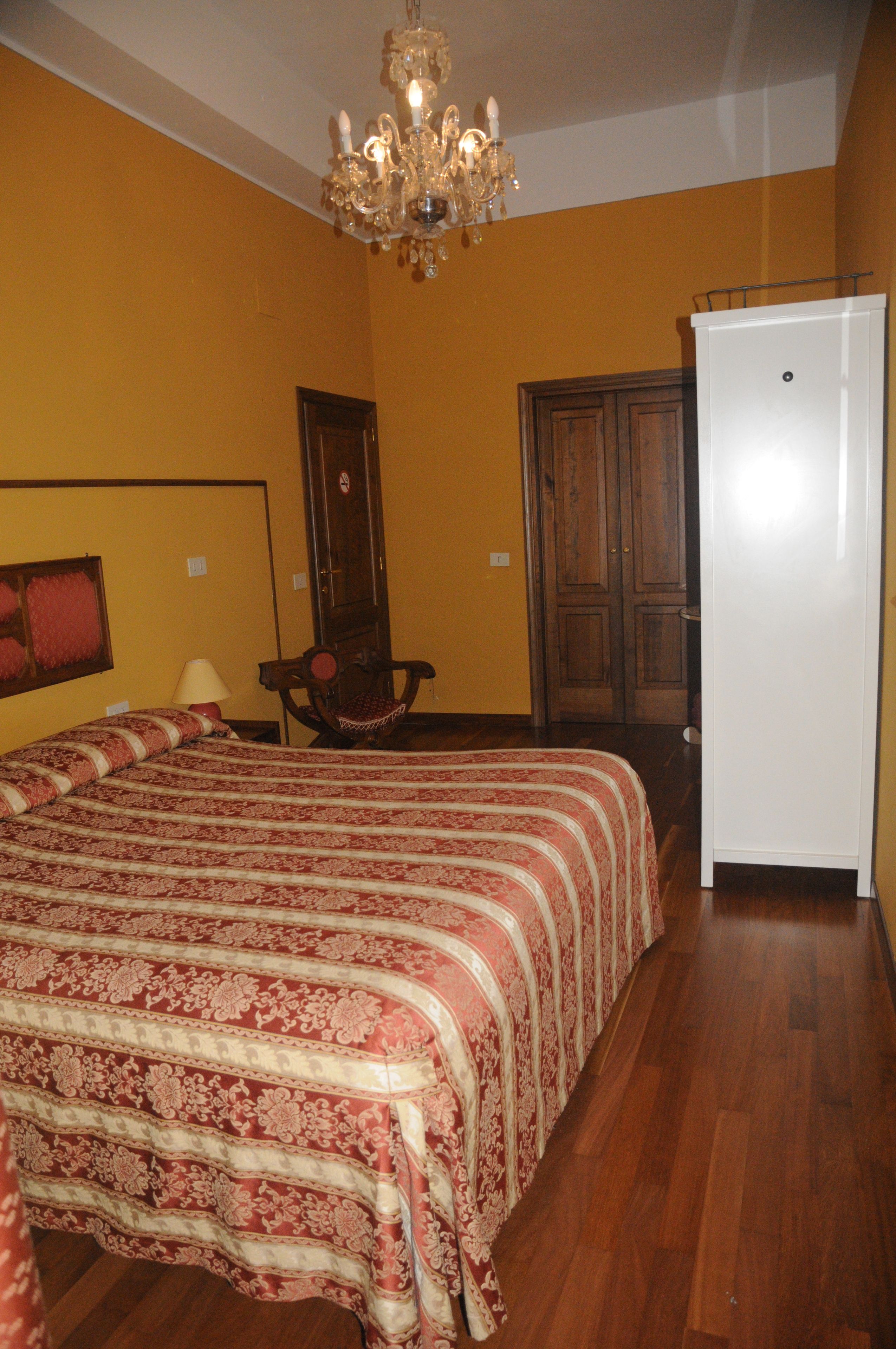 A bedroom with a large bed, a chandelier, wooden flooring, a wardrobe, and a white refrigerator.