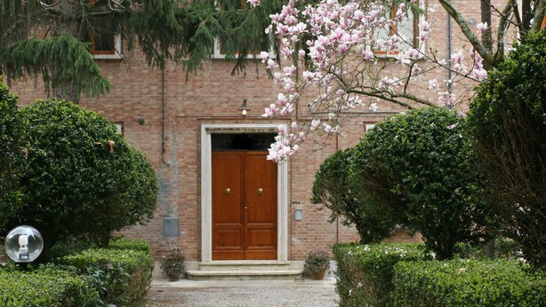 A brick building with a wooden double door, surrounded by well-maintained hedges and flowering trees. - La Coperta Ricamata (Siena)