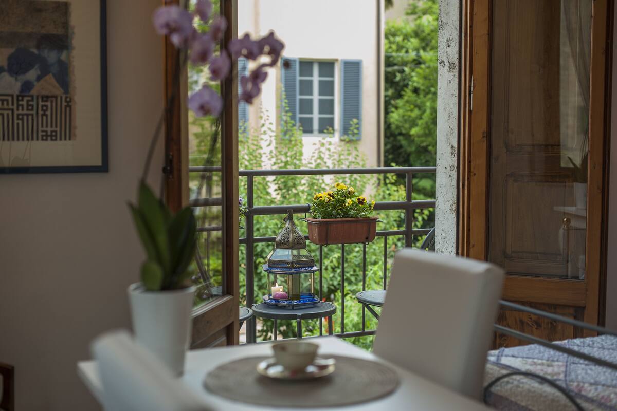 Un balcone con un tavolo, sedie, una pianta in vaso, un portavaso sospeso con fiori e una lanterna decorativa.
