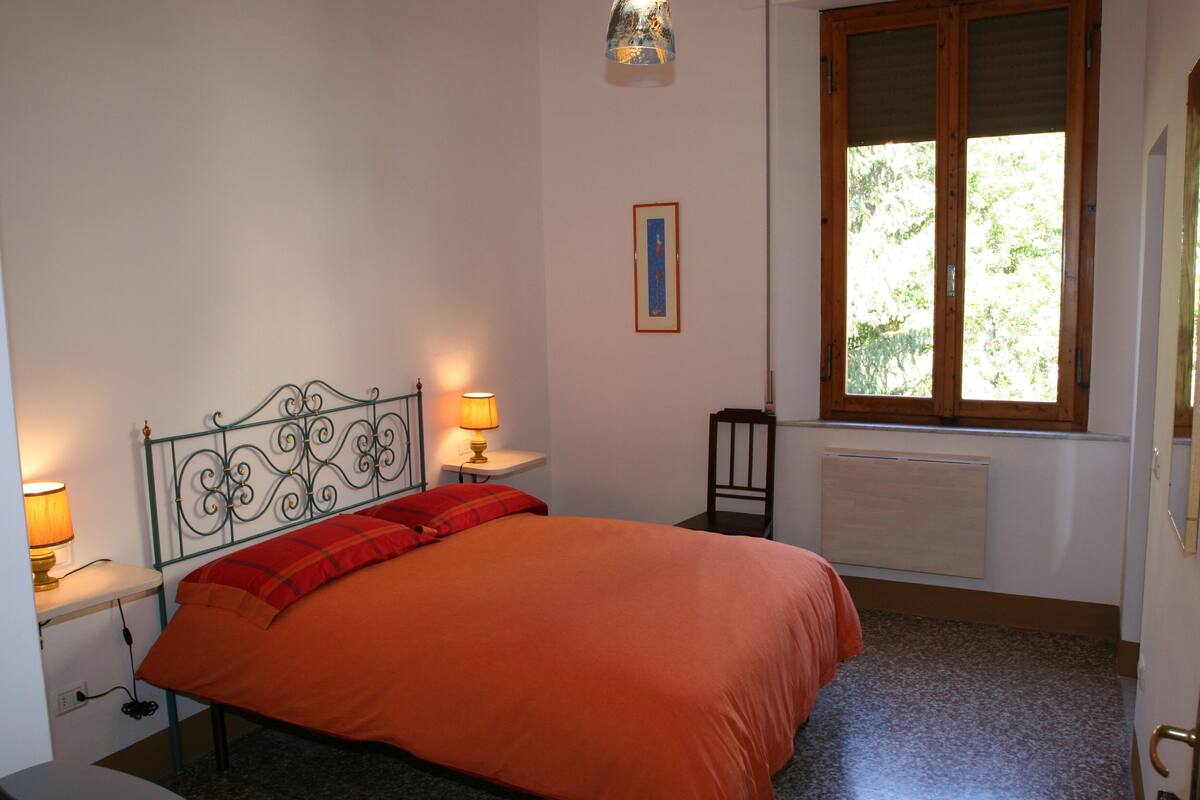 Una camera da letto con testiera in metallo, copripiumino arancione, due lampade sui comodini, una finestra con persiane e un termosifone.
