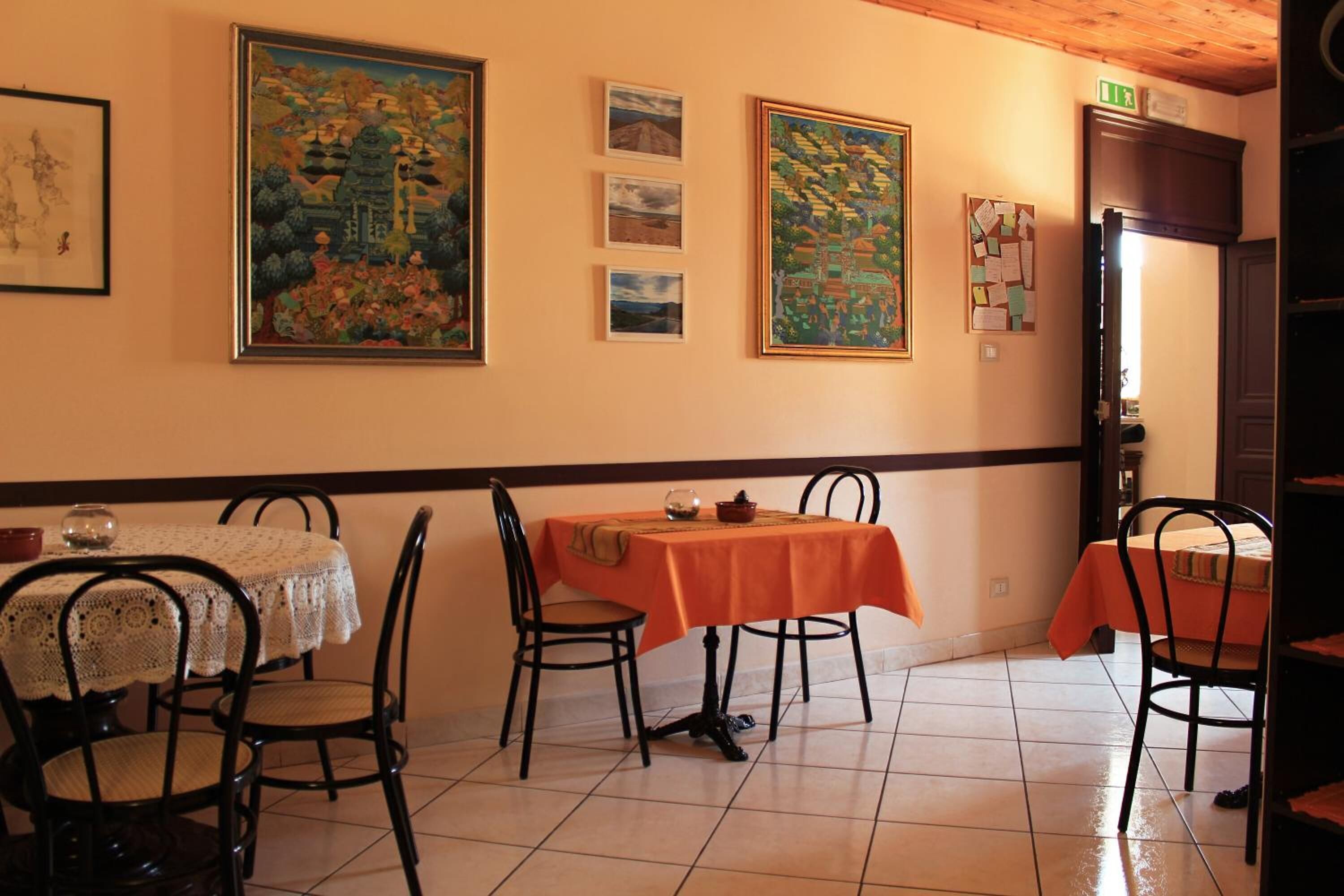 A dining area with two tables, one with a lace tablecloth and one with an orange tablecloth. There are framed artworks on the wall and a bulletin board with papers.