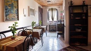 A dining area with a wooden ceiling, tiled floor, and a table set with a tablecloth and chairs. There are potted plants on the table and a bookshelf with various items on the right. - Case D'Anna (Castellammare del Golfo)