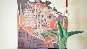 A framed cityscape artwork with a map and text, a vase with white flowers, and a small candle holder on a round table. - Case D'Anna (Castellammare del Golfo)