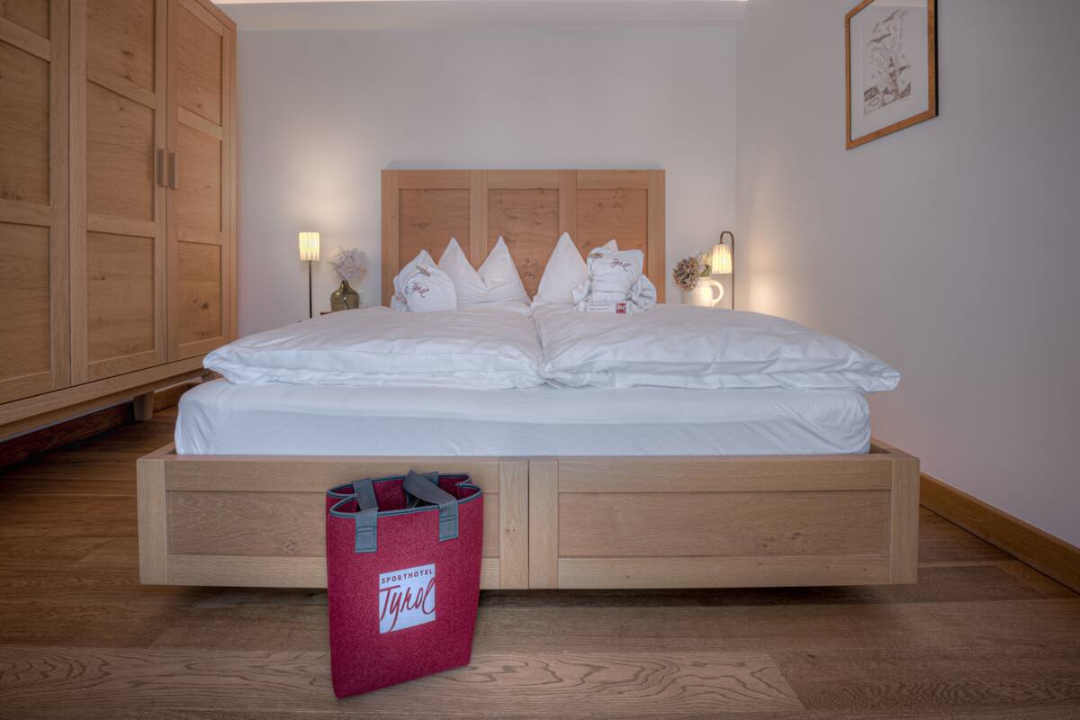 Een netjes opgemaakt bed met witte lakens en kussens, een houten hoofdbord en een rode tas met de tekst 'SPORTHOTEL TYROL' op de vloer.
