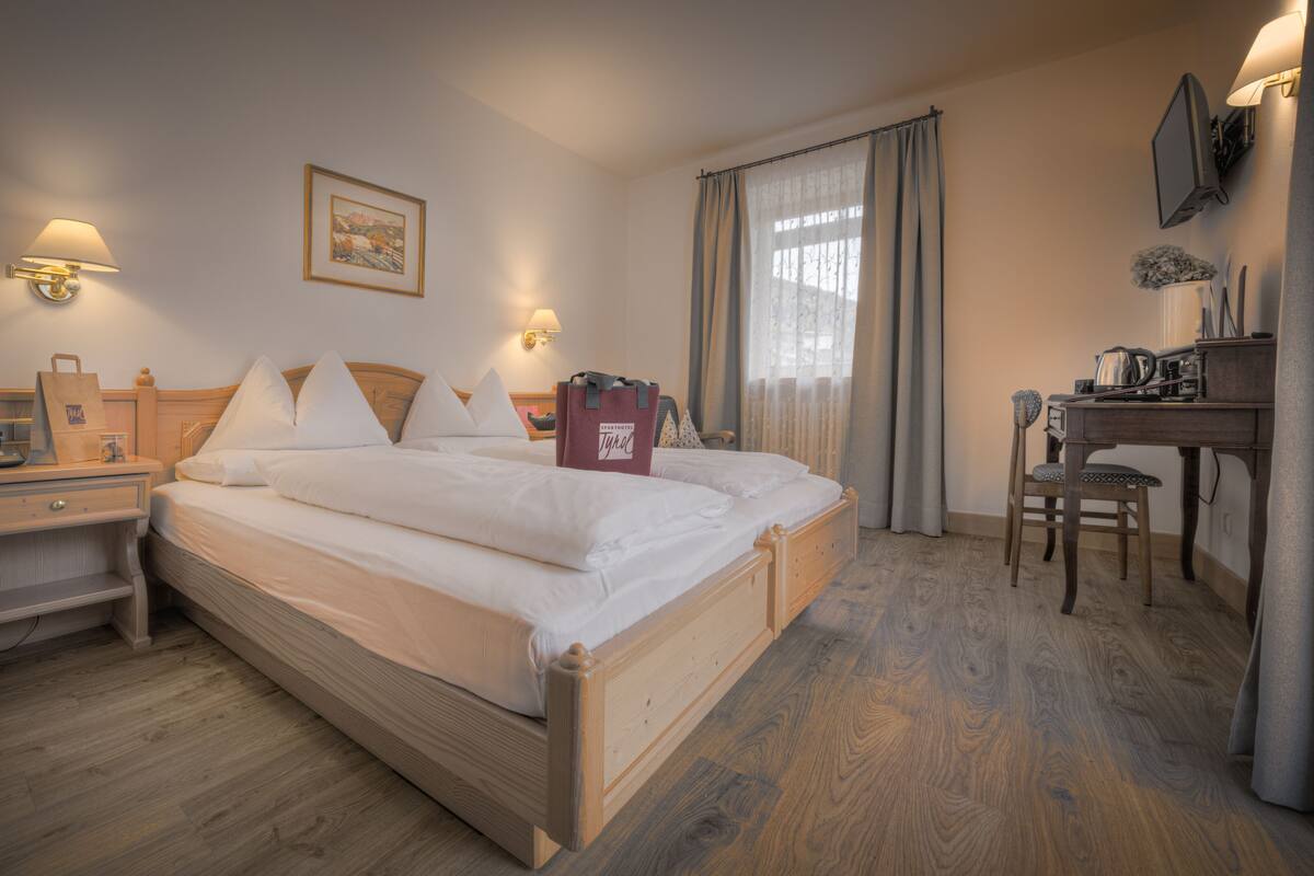 Een hotelkamer met een houten bed, nachtkastjes, een bureau, een stoel, een televisie en een raam met gordijnen.