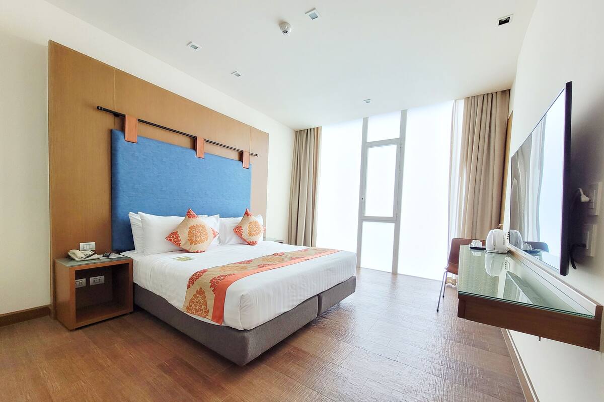A hotel room with a large bed, a nightstand with a phone, a desk with a computer, and a large window.