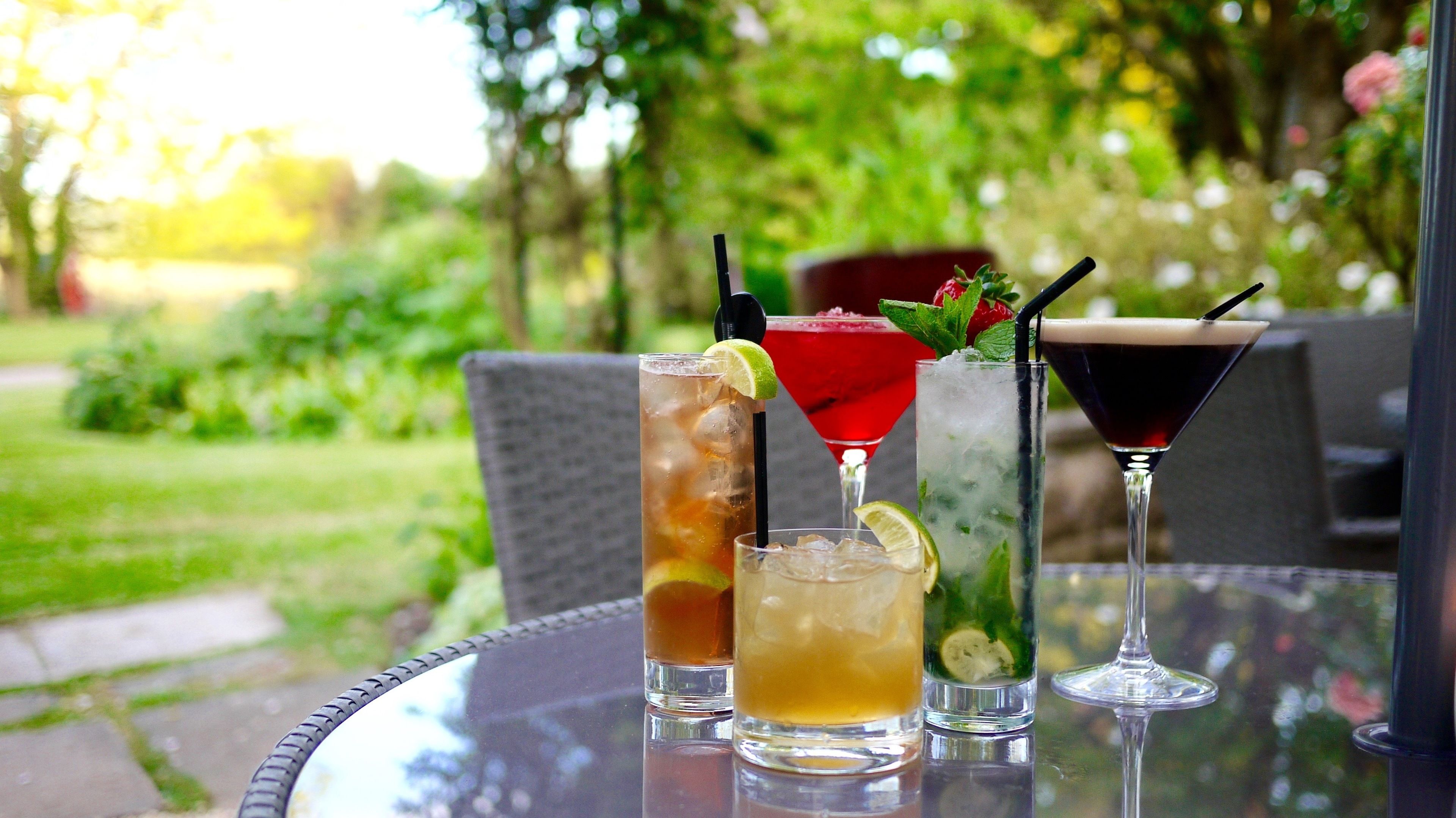 Una mesa con tres bebidas: té helado en un vaso, un cóctel rojo y una bebida oscura, ubicada al aire libre con vista a un jardín.
