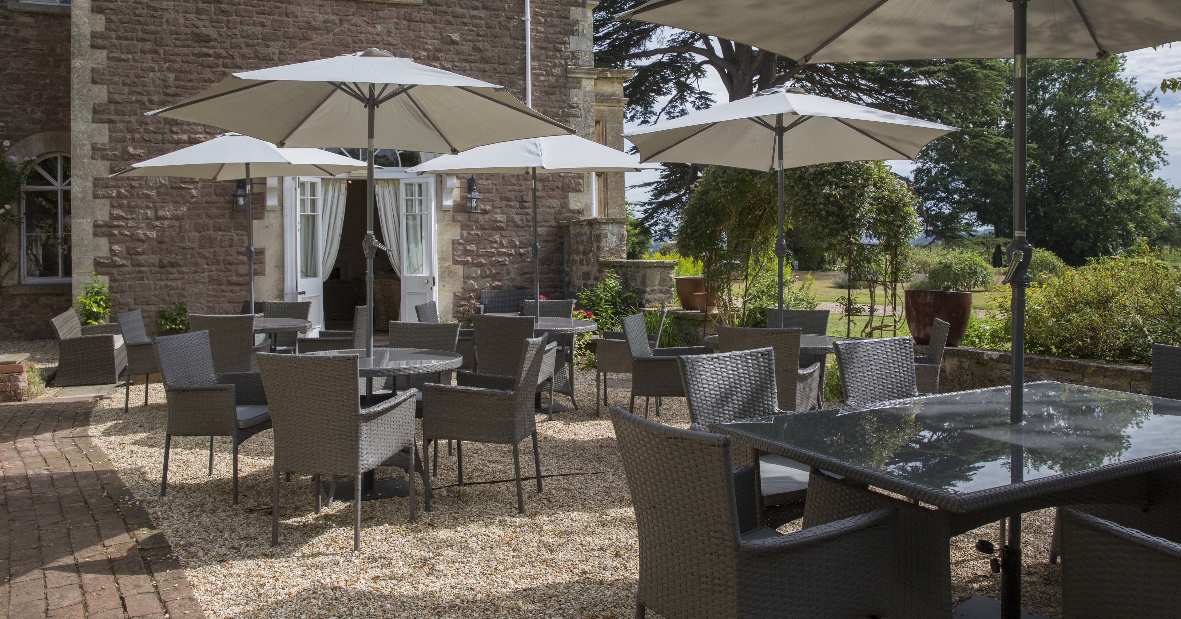 Une terrasse extérieure aménagée avec des meubles en osier et de grands parasols, située devant un bâtiment en pierre donnant sur un jardin.