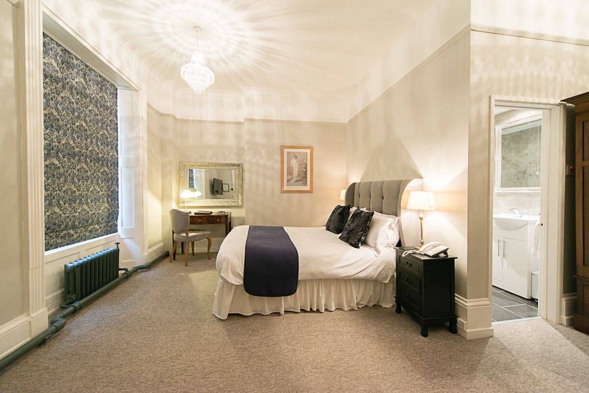 Une chambre à coucher avec un lit, une table de chevet, une lampe et une commode. On y trouve également un bureau avec une chaise et un miroir. Le sol est recouvert d’une moquette et la pièce est équipée d’un radiateur.