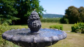 Une fontaine en pierre dont l’eau jaillit du centre, entourée d’une pelouse et d’arbres.