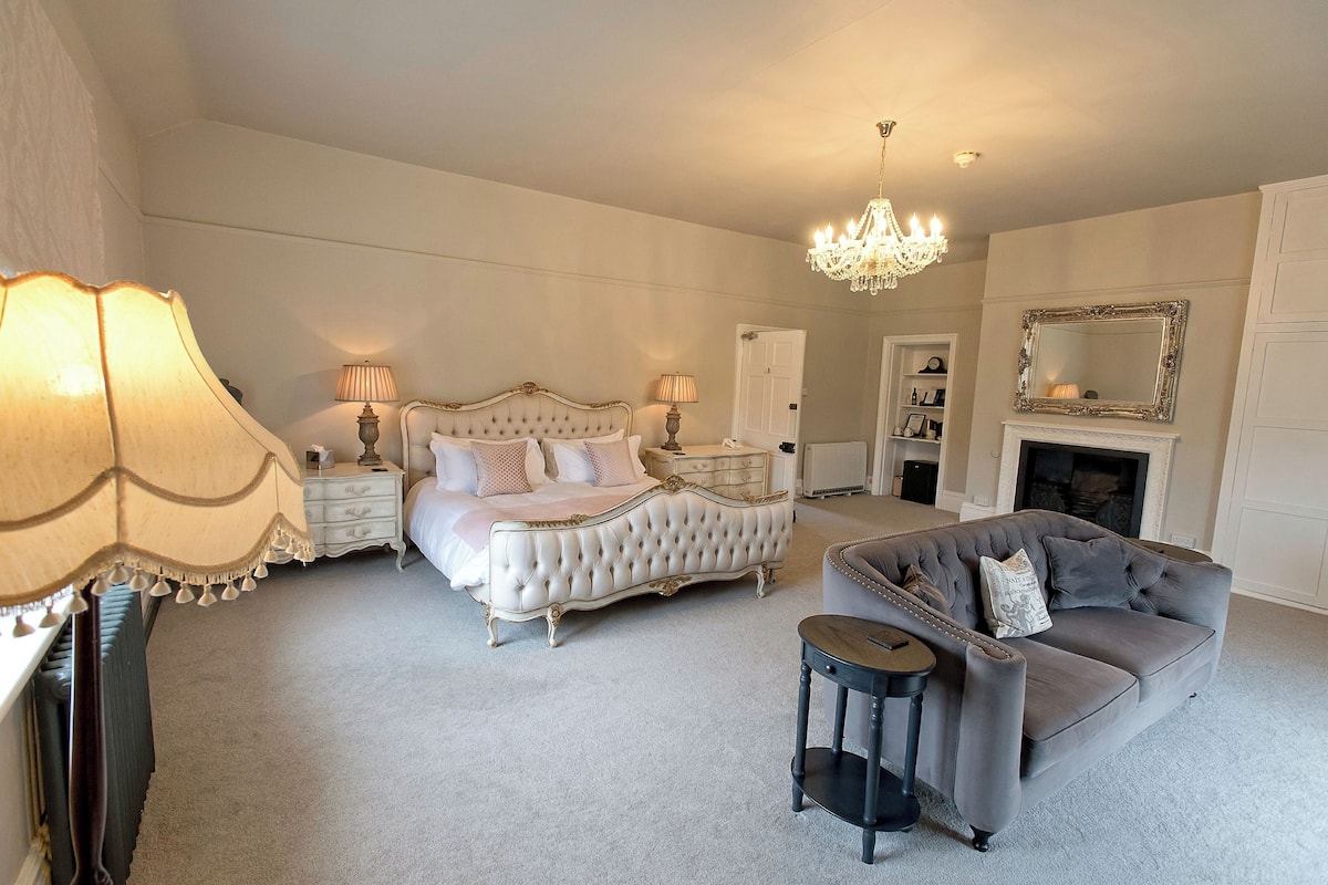 Une chambre avec un grand lit, un canapé gris, un lustre et une cheminée.