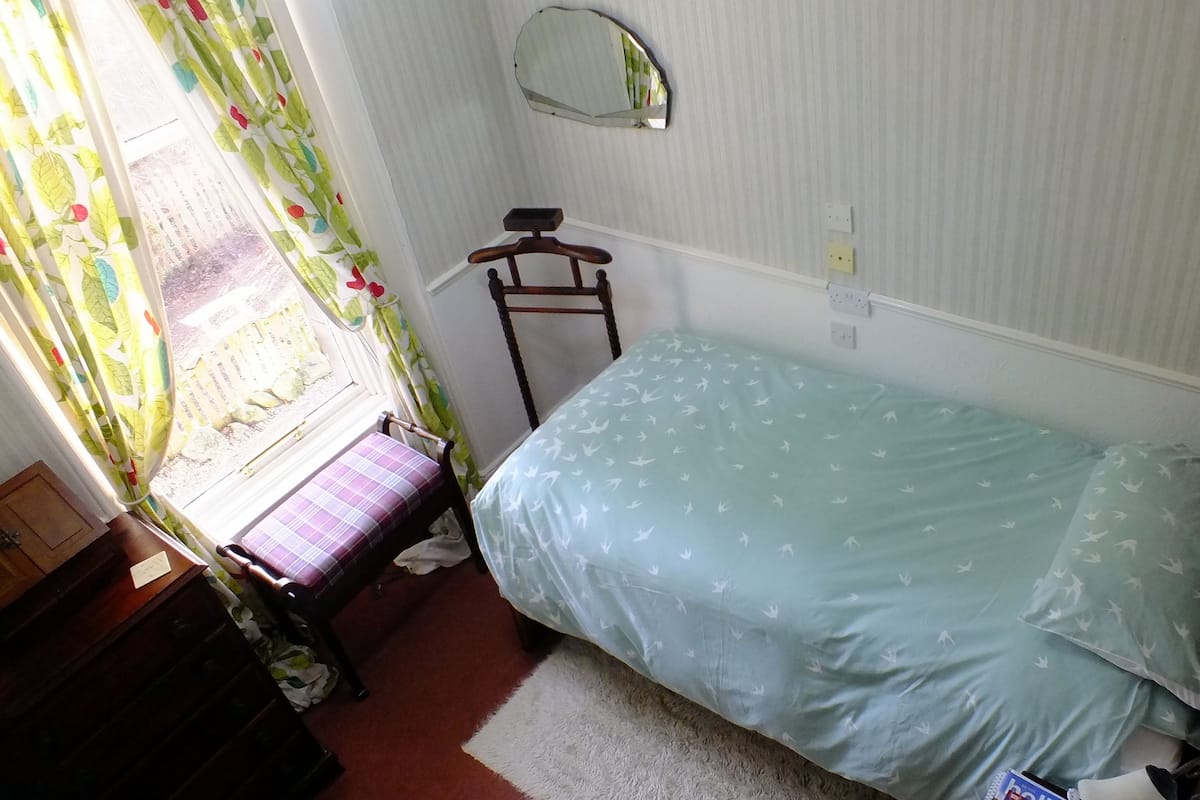 A small bedroom with a single bed, a wooden chair, a mirror, and a window with curtains.