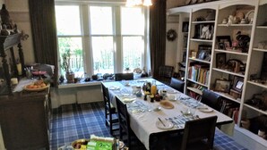 A dining area with a table set for breakfast, a bookshelf with various items, and a window with curtains. - Greystones B&B (Kingussie)