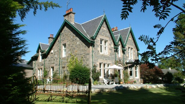 A stone building with a green roof and multiple chimneys, surrounded by a well-maintained garden and outdoor seating area. - Greystones B&B (Kingussie)