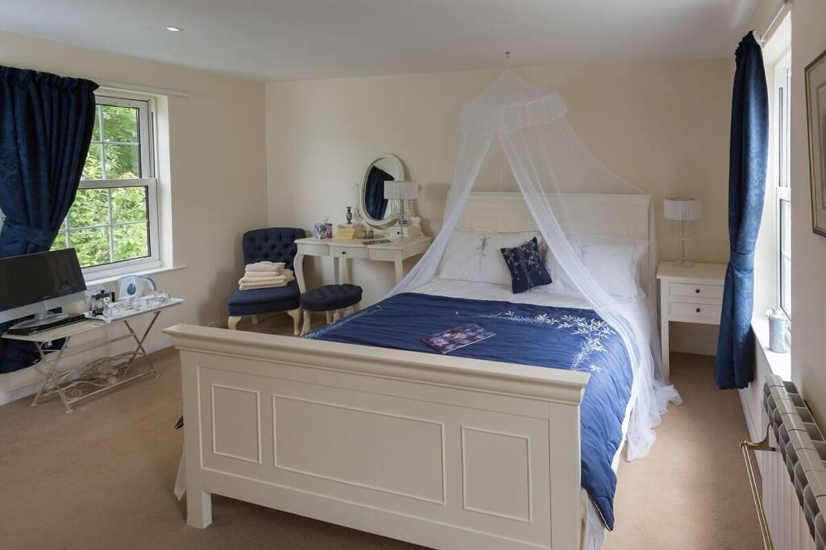 A bedroom with a white four-poster bed, a desk with a chair, a vanity with a mirror, and a window with curtains.