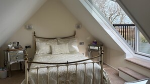 A bedroom with a sloped ceiling, a bed with a metal frame, a chandelier, bedside tables, and a window with a view of the outside. - Clos De Vaul Creux (Sark)