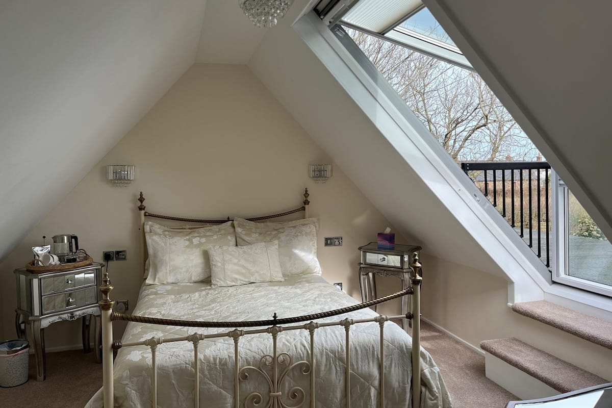 A bedroom with a sloped ceiling, a bed with a metal frame, a chandelier, bedside tables, and a window with a view of the outside.