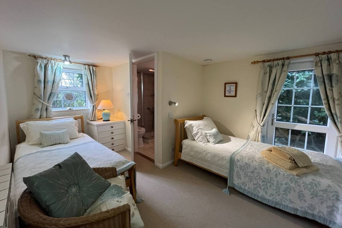 A bedroom with two beds, a bedside table with a lamp, a window with curtains, and a door leading to a bathroom.