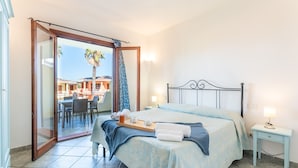 A bedroom with a bed, a bedside table, a nightstand, a window with curtains, and a view of an outdoor area with tables and chairs. - Palau Green Village (Palau)