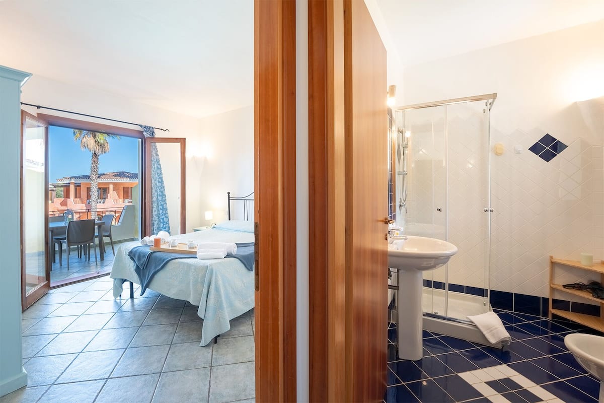 A hotel room with a balcony, a bed, a bathroom with a sink and shower, and a dining area with chairs and a table.