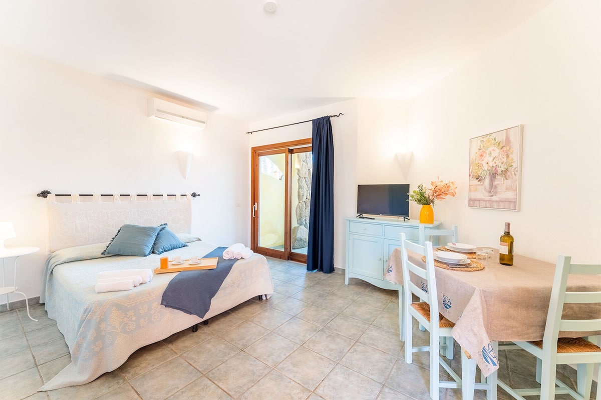 A bedroom with a bed, bedside table, and a TV. There is a dining area with a table and chairs.