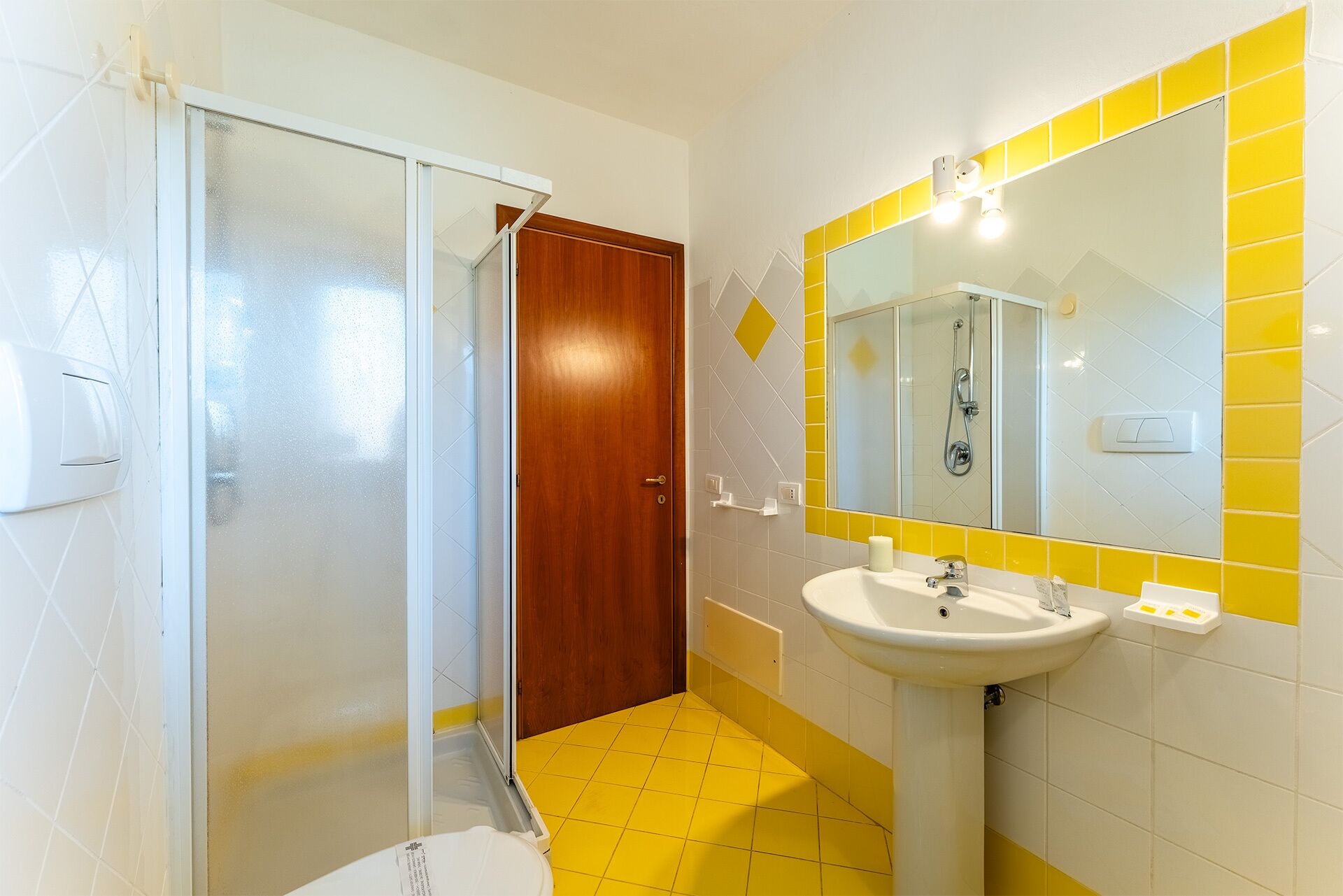 Ein Badezimmer mit Duschabtrennung aus Glas, einer Holztür, einem Waschbecken mit Spiegel und Wandfliesen in Gelb und Weiß.