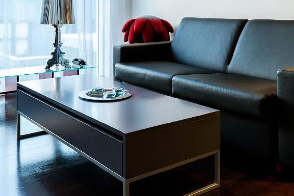 Uma sala de estar moderna com um sofá de couro escuro, uma mesa de centro de vidro e uma almofada vermelha. Há um quadro na parede e um abajur sobre uma mesinha lateral.