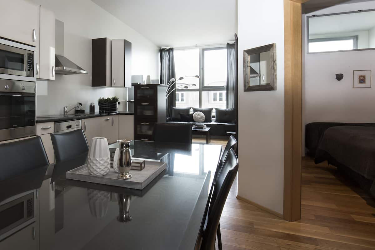 Uma cozinha moderna com eletrodomésticos de aço inoxidável, uma sala de jantar com mesa de vidro e vista para o horizonte urbano através de grandes janelas.