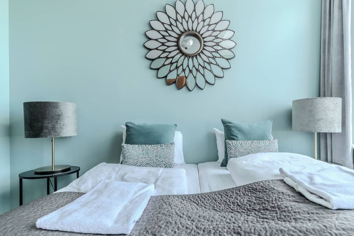 Cama bem arrumada com roupa de cama branca e cinza, duas mesas de cabeceira com luminárias e um objeto decorativo na parede.