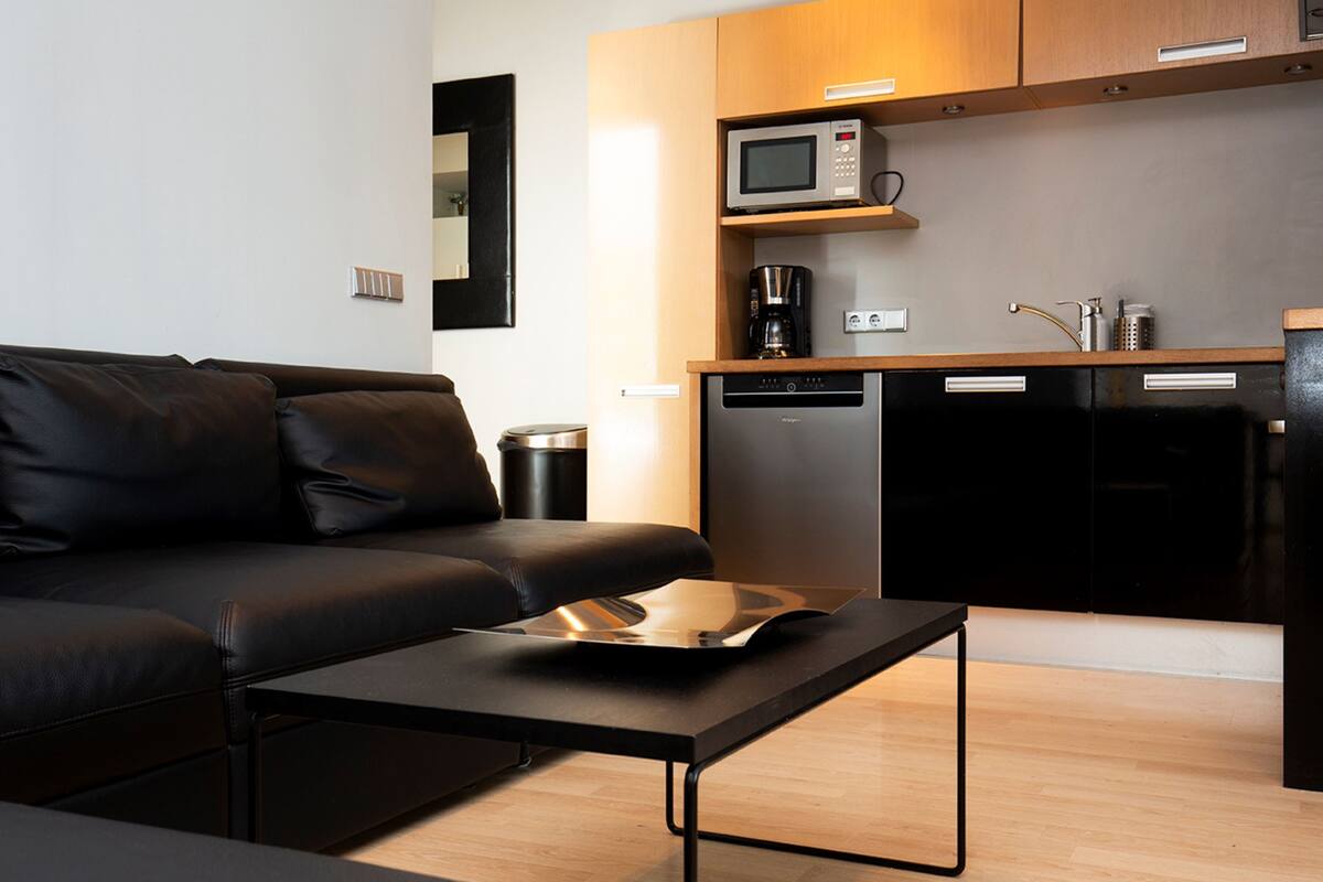 Uma sala de estar moderna com um sofá de couro preto, uma mesa de centro preta e uma área de cozinha com micro-ondas, cafeteira e lava-louças.